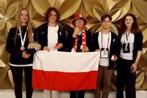 Uśmiechnieta grypa młodzieży z flagą polski przed sobą i madalami w ręka, jedna osoba ma medal w zębach, w tle złota elewacja.