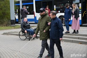 Mężczyzna z opaską na oczach i laską osoby niedowidzącej prowadzony przez drugą osobę