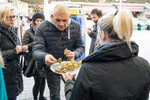 Mężczyzna w średnim wieku sięga po kanapkę do talerza, który trzyma w ręku kobieta podczas degustacji na Halach targowych. Za mężczyzną stoją trzy inne osoby uczestniczące w degustacji.