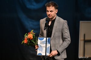 Aktor Teatru Muzycznego Jakub Badurka w eleganckim stroju po odebraniu Nagrody Prezydent Miasta Gdyni z okazji Międzynarodowego Dnia Teatru. W rękach trzyma kwiaty oraz dyplom 