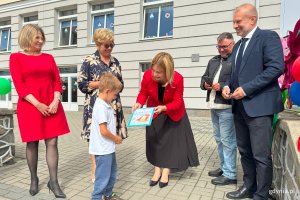 Chłopiec wręcza kobiecie ubranej na czerwono obrazek wykonany z wycinanek, do okoła przed zejściem po schodach stoją dorośli i usnmiechają sie 