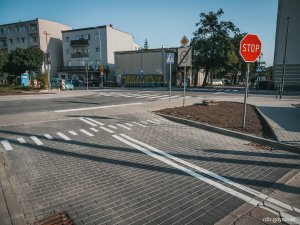 chodniki, wyniesione skrzyżowanie z kostki oraz przejścia dla pieszych po przebudowie. Widoczne oznakowanie, w tle budynki i tereny zielone