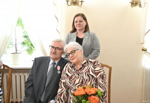 Para starszych ludzi na krzesłach. Za nimi stoi kobieta w szarej marynarce