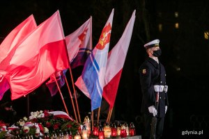 Marynarz stojący przy pomniku Ofiar Grudnia '70 na placu Wolnej Polski podczas uroczystości 50. rocznicy wydarzeń grudniowych. Za nim powiewają flagi narodowe i gdyńskie. Na dole, na płycie pomnika palą się różnokolorowe znicze // fot. Karol Stańczak