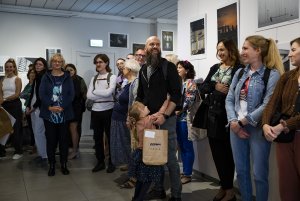 Uśmiechnięty mężczyzna z dzieckiem na gali nagród w galerii fotografii, wokół tłum ludzi.