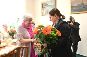 Kobieta w marynarce wręcza bukiet pomarańczowych kwiat&oacute;w staszej pani