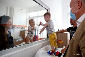 W pokoju szpitalnym stoi prezydent Gdyni, mama małego dziecka i tata innego dziecka. Dzieci witają się przez oddzielającą je szybę.