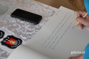Kobieta dokonująca długopisem wpisu w otwartej księdze leżącej na stole z białym obrusem
