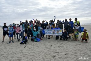 Uczestnicy akcji  „Bałtycka Odyseja”, wraz z wiceprezydentem Gdyni Michałem Guciem, na gdyńskiej plaży. Osoby z przodu trzymają baner z nazwą akcji. Pozostali machają i w rękach trzymają worki oraz chwytaki // fot. Magdalena Czernek