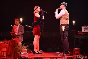 Hala Polsat Plus Arena Gdynia, scena, aktorzy na scenie, aktorka i aktor śpiewają do mikrofonu, dekoracje, czerwone światło, ciemność w tle