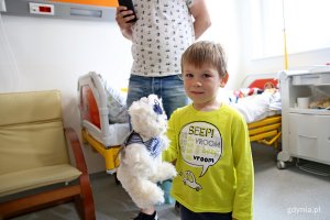 W pokju szpitalnym stoi chłopiec z misiem pluszowym w ręku.