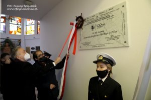 Prezydent Gdyni Wojciech Szczurek oraz Marek Gróbarczyk - sekretarz stanu w Ministerstwie Infrastruktury odsłaniają biała tablicę pamiątkową z czarnymi napisami przygotowaną na stulecie Uniwersytetu Morskiego w kościele oo. Redemptorystów w Gdyni. Na pierwszym planie studentka w mundurze, czarnej maseczce i z białą czapką na głowie // fot. Cezary Spigarski