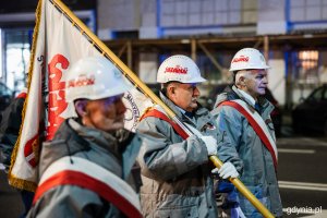 Trzej mężczyźni w kaskach, biało-czerwonych szarfach i ze sztandarem NSZZ „Solidarność”