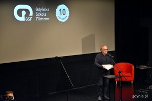 Robert Gliński - reżyser i scenarzysta wygłaszający wykład inauguracyjny z okazji nowego roku akademickiego w Gdyńskiej Szkole Filmowej. W tle wyświetla się logo Gdyńskiej Szkoły Filmowej, po prawej stoi czerwone krzesło // fot. Magdalena Czernek