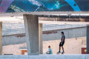 Mężczyzna jadący na deskorolce pod estakadą i mijający drugiego, siedzącego mężczyznę
