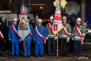 Stoczniowcy ze sztandarami stoją w rzędzie.