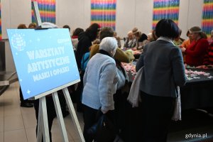 Pomieszczenie w Polsat Plus Arenie Gdynia, tablica z napisem "Warsztaty artystyczne - maski i opaski", grupa starszych osób przy stole z kolorowymi ozdobami