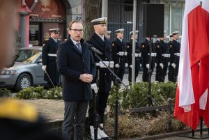 Jakub Ubych - wiceprzewodniczący Rady Miasta Gdyni wygłosił okolicznościowe przemówienie z okazji Narodowego Dnia Pamięci Żołnierzy Wyklętych. Za nim stoją marynarze w galowych mundurach. Po prawej stronie zdjęcia: biało-czerwone flagi // fot. Kamil Złoch