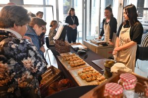 Z lewej strony panie przyglądające się produktom do degustacji, na środku kanapki z różnymi produktami, ułożone na deskach na ladzie, za ladą panie w kuchennych fartuszkach