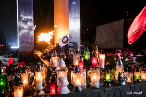 Różnokolorowe znicze ustawione na pomniku Ofiar Grudnia '70 na placu Wolnej Polski w Gdyni. Przy jednym z nich zatknięto białą flagę z czarnym logiem Grudnia 1970. W tle płonącą na pomniku pochodnia // fot. Karol Stańczak