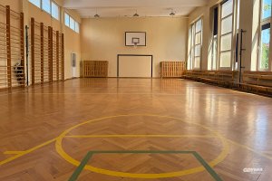 Parkiet z liniami do gier zespołowych, na ścianie po lewej stronie drabinki do ćwiczeń, na końcy sali bramka i tablica do koszykówki powyżej
