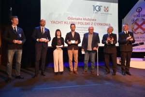 Grupa osób stojących na scenie i mających za plecami biały ekran