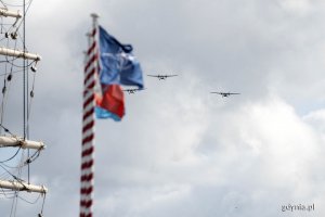 Na pierwszym planie maszty z powiewającymi flagami NATO i Polski. Z nimi na niebie widać samoloty transportowe przelatujące nad skwerem Kościuszki podczas pokazu z okazji święta 43. Bazy Lotnictwa Morskiego i Dywizjonu Okrętów Bojowych 
