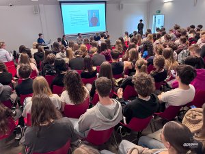 Ujęcie z tyłu młodzieży siedzącej w sali konferencyjnej, w tle kilka osób opowiadających o swoich doświadczeniach na emigracji.