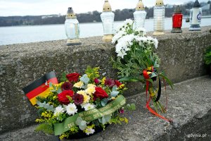 Wieniec i wiązanka kwiatów oraz znicze ułożone na nabrzeżu przy molo Południowym