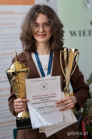 dziewczyna z kręconymi włosami do ramion i w okularach, ubrana w brązowa marynarkę, trzymająca 2 puchary oraz dyplomy 