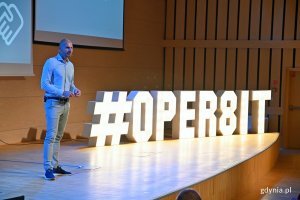 Krzysztof Kozakiewicz, organizator konferencji i CEO fundacji Let's Manage IT, podczas wystąpienia na konferencji OPER8IT. Po prawej: podświetlony napis #OPER8IT // fot. Magdalena Czernek