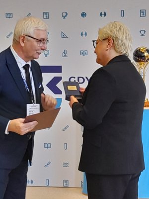 Z lewej strony profesor stoi bokiem do obiektywu, Kurator stoi tyłem do obiektywu trzyma w ręcę odznaczenie wręczone przez pana Profesora 
