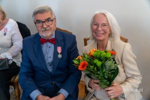 Mężczyzna w garniturze, z medalem w klapie i uśmiechanięta kobieta z kwiatami
