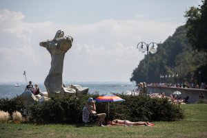 Pomnik oraz ludzie opalający się na trawie pod parasolem. W tle część bulwaru. 