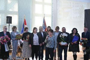 Stojąca kobieta mówiąca do mikrofonu w otoczeniu innych osób w szkolnej hali sportowej