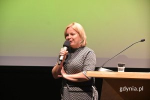 Kobieta stojąca na scenie obok mównicy i mówiąca do mikrofonu trzymanego w ręce