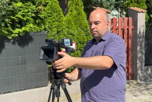 Upalny dzień, mężczyzna przygotowuje kamerę video do nagrania, w tle ogrodzenie i kawałek zieleni