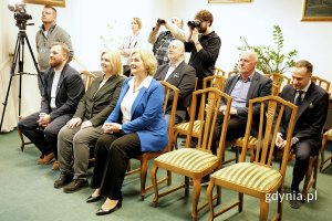 Grupa osób siedzących na krzesłach w eleganckiej sali, w tle widoczni fotoreporterzy