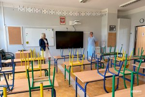 Pomieszczenie z ławkami szkolnymi i krzesłami postawionymi do góry nogamia, w tke dwie osoby tablica szkolna