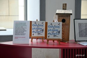 Dwa egzemplarze książki „Gdynia: ptasie miasto” oraz budka lęgowa na stoliku w holu Muzeum Miasta Gdyni. Po lewej - informacje, że dochód ze sprzedaży książki pokrył zakup i montaż wyklejki z markerami na szybach muzeum