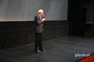 Mężczyzna w garniturze z mikrofonem na scenie na tle kinowego ekranu