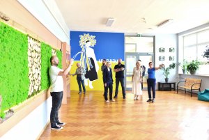 Grupa osób w holu szkoły, na ścianie mural w barwach Ukrainy z kobiecą postacią, po lewej stronie zielona ściana z chrobotka, ramy drewniane, po prawej okna, dużo kwiatów w doniczkach