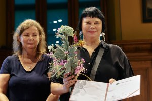 dwie kobiety, uśmiechnięta trzyma kwiaty i dyplom