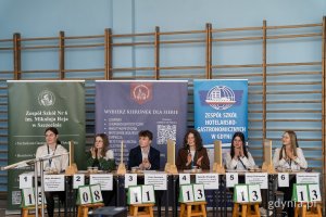 sześcioro uczestników konkursu, siedzą przy biurkach, w tle banery promocyjne i sala gimnastyczna 