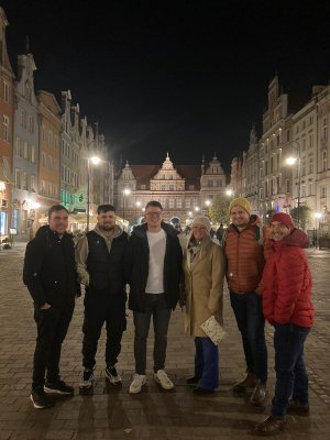 Na zdjęciu znajduje się grupa uczetsników stażu międzynarodowego, ubrani w kolorową odzień. Występują na tle zabytków Gdańska, wieczorową porą.