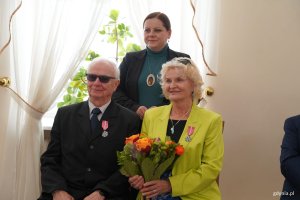 Para siedzi, kobieta trzyma pomarańczowe kwiaty, z tyłu stoi inna kobieta
