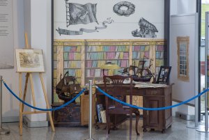 Fragment wystawy  „Akwarela, pastel, piórko – w służbie mistrza ilustracji Eugeniusza Koczorowskiego”: drewniane biurko, na którym stoją: lampa, model galeonu i zdjęcie kmdr. Eugeniusza Koczorowskiego. Całość na tle ścianek, na których namalowano regał z książkami i okno // fot. materiały MMW