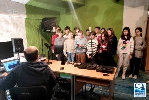 W dalszym planie dzieci w studio podczas realizacji nagrań audio, na pierwszym planie nauczyciel odwrócony do nich twarzą 