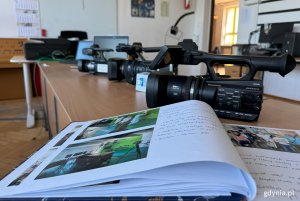 Sala ze stołami, na których stoją kamery video, na pierszym planie kronika, w dalszym planie radiowęzeł i drzwi wyjściowe z sali