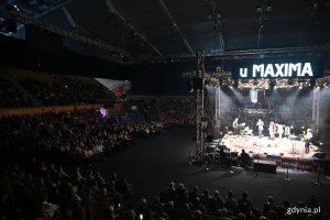 Hala Polsat Plus Arena Gdynia, scena, dekoracja ze stołem i krzesłami, aktorzy na scenie, publiczność dookoła, nad sceną wyświetlacz z napisem "U Maxima"